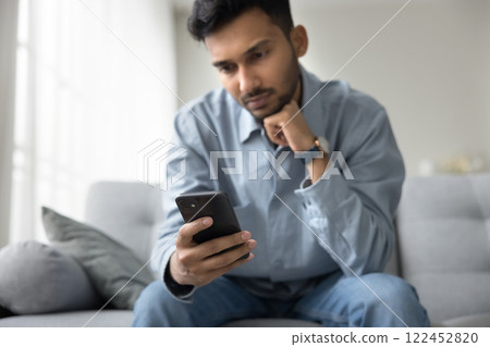 Serious millennial male sit on sofa read news on smartphone 122452820