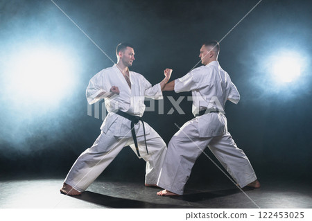 Martial arts masters on dark background. Strong man fighters in white kimono for karate martial arts fighting training in black gym. Karate fighters training punches kicks practicing fight sparring Martial arts masters on dark background. Strong man fighters in white kimono for karate martial arts fighting training in black gym. Karate fighters training punches kicks practicing fight sparring 122453025