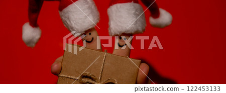 Fingers dressed in Santa-Claus red-white hats. Two happy finger smileys faces holding paper gift box present against red background. Happy family celebrating concept for Christmas or New Years day Fingers dressed in Santa-Claus red-white hats. Two happy finger smileys faces holding paper gift box present against red background. Happy family celebrating concept for Christmas or New Years day 122453133