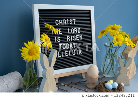 Phrase CHRIST THE LORD IS RISEN TODAY ALLELUIA traditional greeting to christian holiday. Festive mood composition with bunnies and yellow flowers on pastel blue background. Sustainable natural Phrase CHRIST THE LORD IS RISEN TODAY ALLELUIA traditional greeting to christian holiday. Festive mood composition with bunnies and yellow flowers on pastel blue background. Sustainable natural 122453211