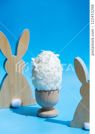 Wooden minimalism low-key bunny decor. Easter egg made of white lilac spring flowers in pastel blue background. Sustainable holiday minimalistic still life composition. Copy space template for 122453266