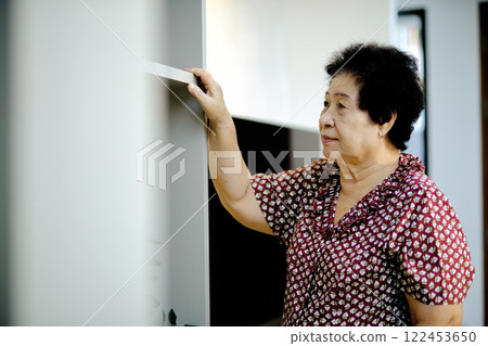 Elderly woman standing near bookshelf, reaching up with one hand, wearing patterned blouse, looking calm and thoughtful indoors 122453650