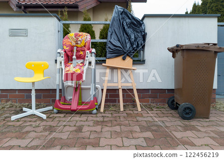 A popular curbside display of unwanted items including a high chair a yellow stool an easel with a trash bag and a brown garbage bin 122456219