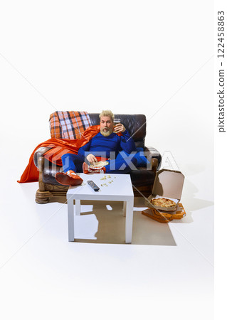 Man in superhero costume sitting on couch, watching TV, eating popcorn and drinking beer, open pizza boxes on floor against white studio background 122458863