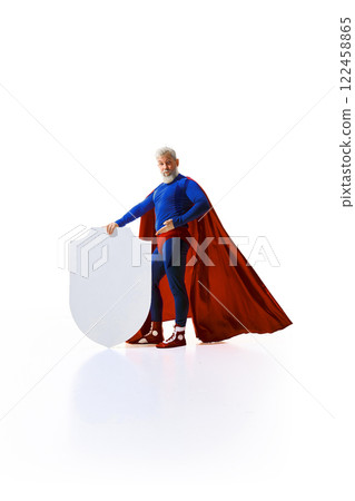 Full-length portrait of confident man in superhero costume with red cape pointing at white shield against white studio background 122458865