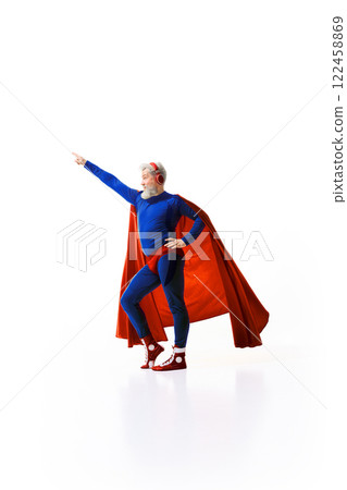 Full-length image of grey-haired man superhero costume with red cape, listening to music in headphones and raising fist up against white studio background 122458869
