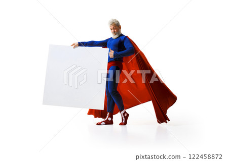 Grey-haired man in superhero costume standing with big white empty poster against white studio background 122458872