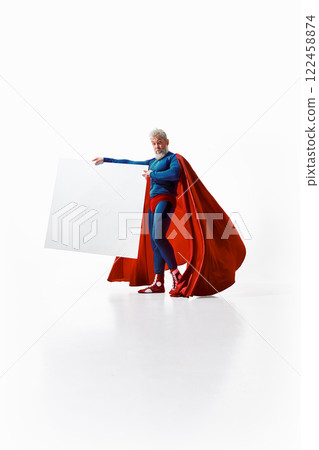 Funny grey-haired man in superhero costume standing in pose of bullfighter with big white poster with empty space against white studio background Funny grey-haired man in superhero costume standing in pose of bullfighter with big white poster with empty space against white studio background 122458874