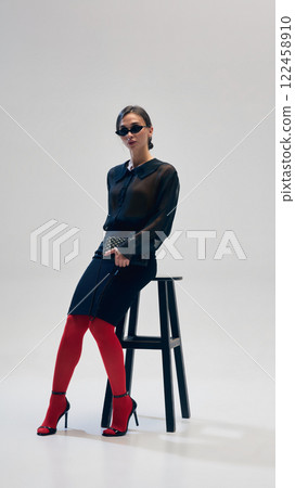 Elegant woman in black blouse, skirt, red tights, heels and sunglasses posing with confidence on chair against grey studio background 122458910