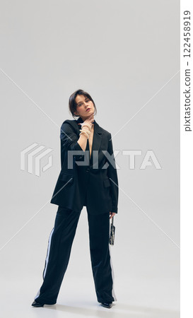 Full-length portrait of beautiful young woman in black suit, pants, blazer, small purse and pearl necklace posing against grey studio background 122458919