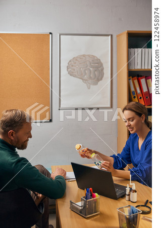 Woman, doctor explaining medication options to man, holding medical bottles, in clinic with organized shelves in the background. 122458974