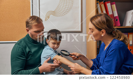 Doctor closely examining young boy arm with magnifying glass, assisted by the his father, in clinical setting. Doctor closely examining young boy arm with magnifying glass, assisted by the his father, in clinical setting. 122458980