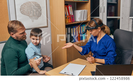 Female doctor explaining health details to father holding young boy during medical consultation, showcasing family-centered pediatric care, in clinical office. Female doctor explaining health details to father holding young boy during medical consultation, showcasing family-centered pediatric care, in clinical office. 122458986