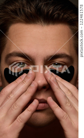 Close up of young man with healthy skin applying moisturized hydrogel black patches against brown studio background. Close up of young man with healthy skin applying moisturized hydrogel black patches against brown studio background. 122461578