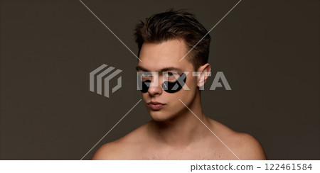 Shirtless young man with black eye patches, looks calm, embodying self-care and anti-fatigue skincare against brown studio background. 122461584