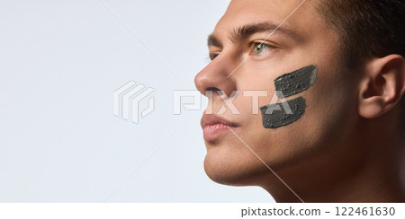 Close up of confident young man with nourishment, moisturized facial clay mask on skin, looking forward against white studio background. 122461630