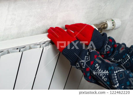 Hands in gloves on a radiator. Central heating radiator at home. A child in woolen gloves warms his hands near the heater Hands in gloves on a radiator. Central heating radiator at home. A child in woolen gloves warms his hands near the heater 122462220