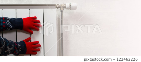 Hands in gloves on a radiator. Central heating radiator at home. A child in woolen gloves warms his hands near the heater Hands in gloves on a radiator. Central heating radiator at home. A child in woolen gloves warms his hands near the heater 122462226