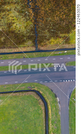 Aerial view of a rural area featuring a straight road with marked lanes, surrounded by green grass, a small canal, and patches of wetland. The vibrant landscape showcases natural and human elements. 122463070