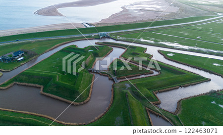 A drone video showing a unique landscape of man-made mounds and waterways near the sea, with a house on the grassy terrain, and a distant view of the coastline stretching under a cloudy sky. 122463071