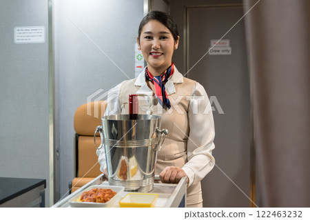 Smiling Asian flight attendant holding food and beverage cart in aircraft cabin for serving passengers 122463232