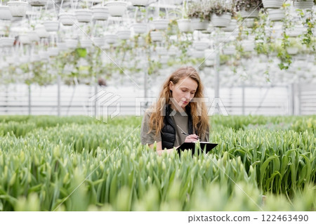 A woman stands in a greenhouse with tulips, watches the growth of flowers and makes notes in a notebook 122463490