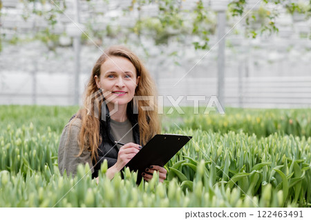A woman stands in a greenhouse with tulips, watches the growth of flowers and makes notes in a notebook 122463491