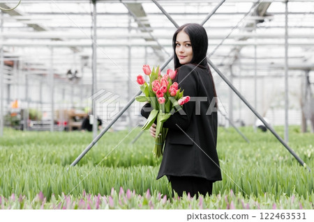 Young woman standing in large greenhouse and holding a blooming tulips in her hands Young woman standing in large greenhouse and holding a blooming tulips in her hands 122463531