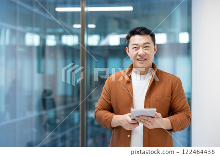 Portrait of successful Asian man inside office. Businessman smiling and looking at camera while holding tablet computer. Programmer developer at workplace near window. 122464438