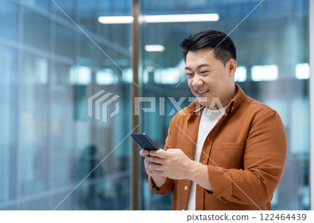 Successful Asian businessman standing near the window with phone in hands. Man using application on smartphone, typing message and browsing social media inside office at workplace. 122464439