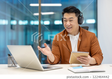 Asian businessman wearing headphones engaging in virtual meeting, holding notebook. Smiling professional in office setting, using laptop for remote work and clear communication, showcasing modern Asian businessman wearing headphones engaging in virtual meeting, holding notebook. Smiling professional in office setting, using laptop for remote work and clear communication, showcasing modern 122464442