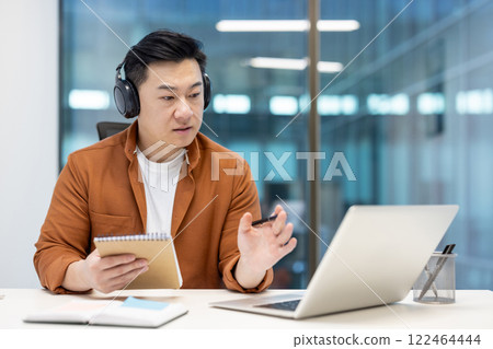 Asian man wearing headphones using a laptop for an online meeting, dressed casually in an office setting. He holds a notepad in one hand and writes with a pen, showing focus and productivity. Asian man wearing headphones using a laptop for an online meeting, dressed casually in an office setting. He holds a notepad in one hand and writes with a pen, showing focus and productivity. 122464444