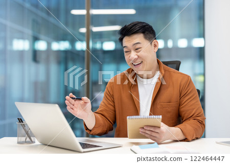 Asian man mature businessman smiling and engaging in an online meeting while taking notes. He is seated in a modern office setting with a laptop and appears happy and professional. 122464447
