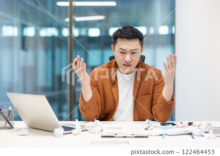 Asian man experiencing frustration and stress while working at a cluttered desk in an office. Overwhelmed by workload, he sits surrounded by crumpled papers and tries to manage challenges. 122464453