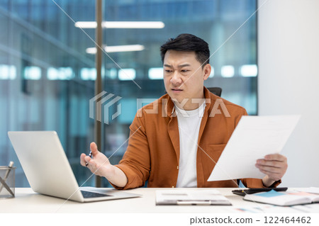 Asian man examining documents with a critical expression while using a laptop in office, reflecting a moment of professional questioning. 122464462