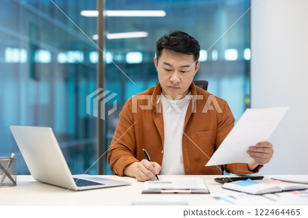 Asian businessman sits at desk working with documents, demonstrating focus and productivity. Man holds pen, works with laptop, showcasing professional business setting. Ideal office themed content. Asian businessman sits at desk working with documents, demonstrating focus and productivity. Man holds pen, works with laptop, showcasing professional business setting. Ideal office themed content. 122464465