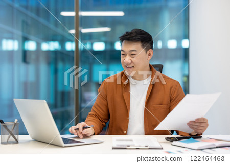 Asian businessman working on laptop holding document, showcasing professional success and focus. Office worker in modern setting reflecting concepts of business, productivity, and technology. 122464468