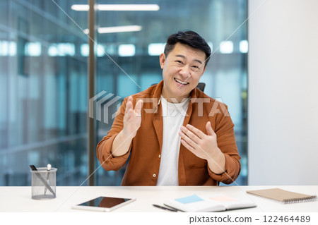 Asian man expressing ideas while seated at a desk in an office setting, conveying confidence, positivity, and communication during a business-related discussion or meeting. 122464489