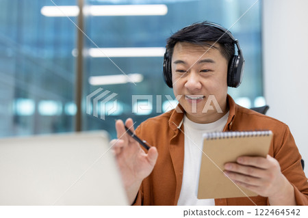 Focused Asian businessman wearing headphones participates in online meeting with notebook and pen in hand. Engaged in remote communication in modern office environment. 122464542