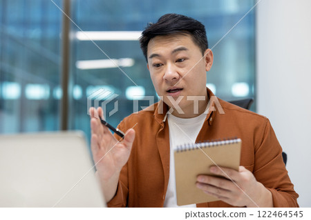 Asian man businessman in office conducting video meeting on laptop, holding notebook and pen, expressing ideas. Bright modern professional setting enhances business communication. 122464545