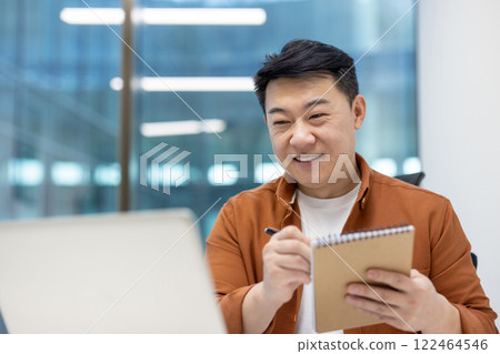 Asian man businessman engaged in business meeting, smiling while taking notes. Modern office setting highlights work environment, illustrating professional engagement, productivity, and positive 122464546
