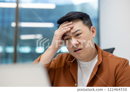 Asian man businessman feeling stressed at work while concentrating on laptop. Office environment with technology and challenges. Business setting showing pressure and problem-solving mindset. 122464550