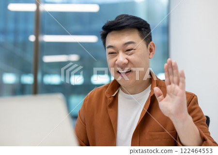 Asian man businessman engaging in virtual meeting, smiling, waving at laptop screen in casual outfit. Modern office setting, focus on communication and technology. Asian man businessman engaging in virtual meeting, smiling, waving at laptop screen in casual outfit. Modern office setting, focus on communication and technology. 122464553