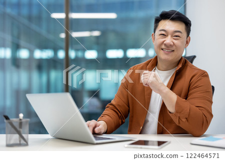Asian man businessman using laptop at office, smiling confidently, demonstrating productivity and business success. Reflects technology usage with phone on desk, symbolizing modern professional life. 122464571