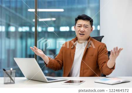Asian man businessman sitting at desk showing confusion or discussion during meeting in modern office setting. Laptop open, phone visible, emotion of confusion or inquiry. Asian man businessman sitting at desk showing confusion or discussion during meeting in modern office setting. Laptop open, phone visible, emotion of confusion or inquiry. 122464572
