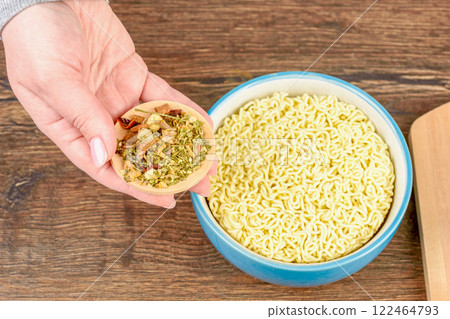 A woman's hand holds a saucer with dry herbs and spices over instant noodles. 122464793