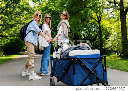 Three young adult beautiful woman girls friends enjoy have fun walking picnic party city park pulling folding cart food drink equipment sunny day. Female person friendship summer outdoors celebration 122464917