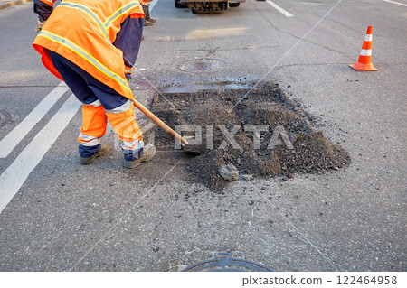 Road workers repair pothole in bright orange uniforms on a busy street. Copy space. Road workers repair pothole in bright orange uniforms on a busy street. Copy space. 122464958