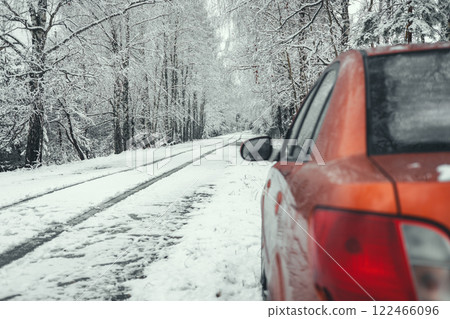 Red car parked along a snowy road winding through a tranquil winter forest, creating a serene atmosphere filled with the beauty of nature and the quiet of the season. 122466096