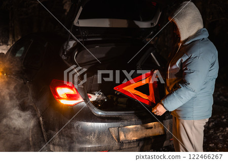 Woman stands by her car on a snowy forest road at night, holding an emergency triangle. Her vehicle's trunk is open, indicating a breakdown in the cold, dark surroundings. 122466267